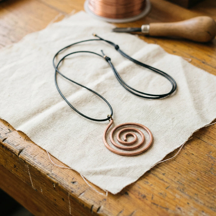 Copper spiral pendant necklace with adjustable black cord resting on linen fabric beside wooden surface
