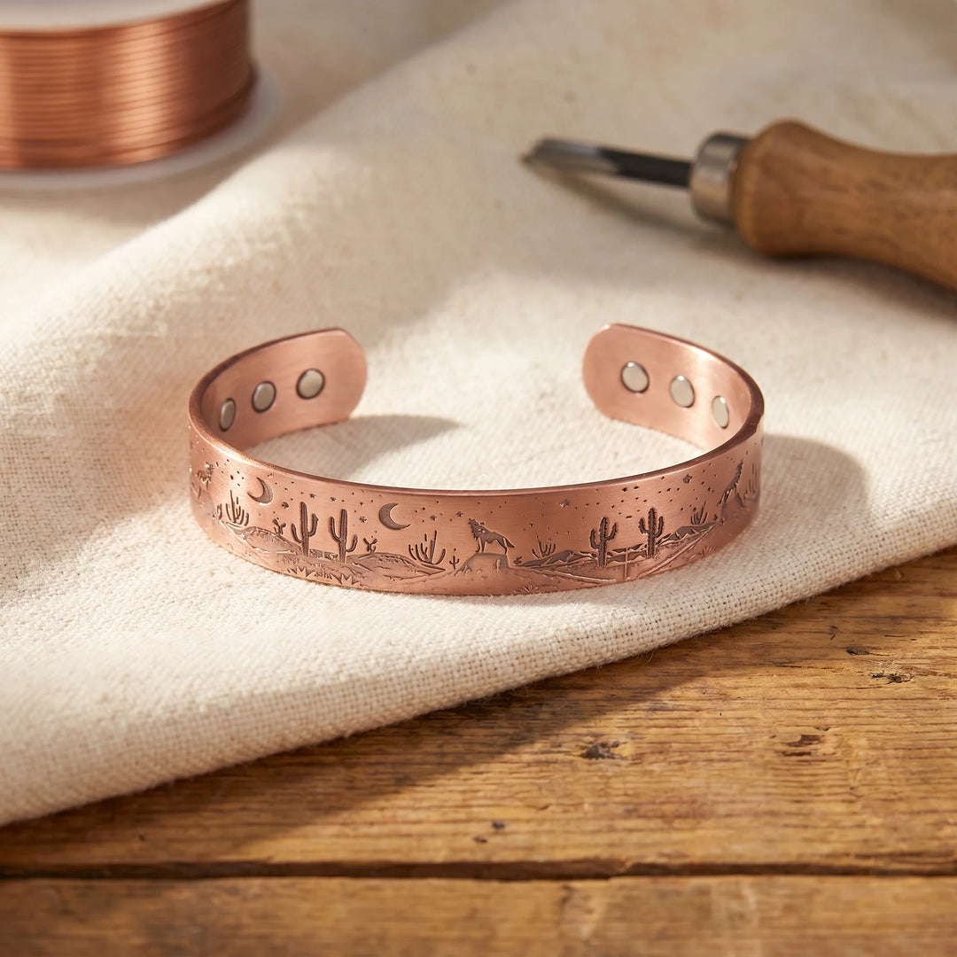 Copper cuff bracelet with engraved desert scene featuring cacti and coyote motif displayed on linen fabric