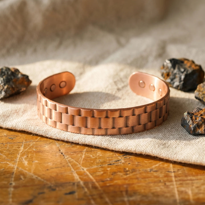Copper cuff bracelet with watch-link texture displayed on linen fabric beside decorative stones