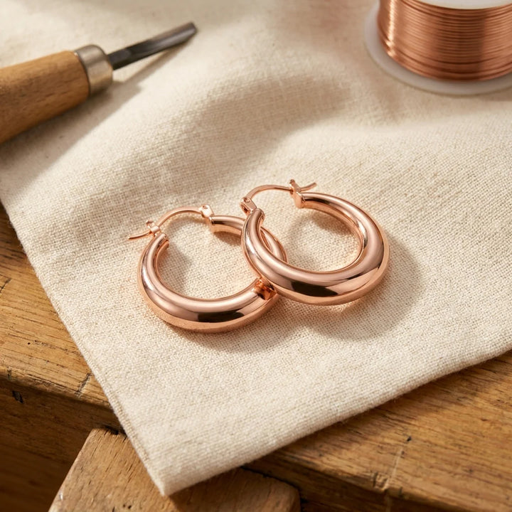 Rose gold hoop earrings with polished finish and hinge clasp resting on neutral linen fabric