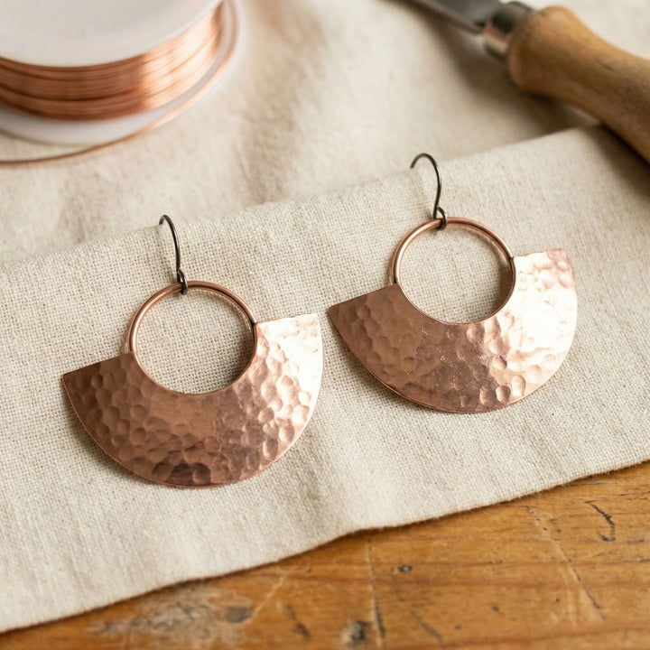 Hammered copper drop earrings with geometric half-circle design displayed on natural linen fabric