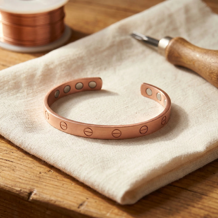 Women's copper cuff bracelet with engraved screw-head motif displayed on linen fabric beside crafting tools