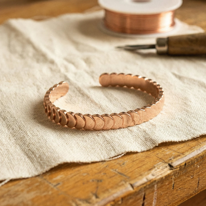 Women's copper cuff bracelet with overlapping scale design resting on linen fabric beside copper wire spool