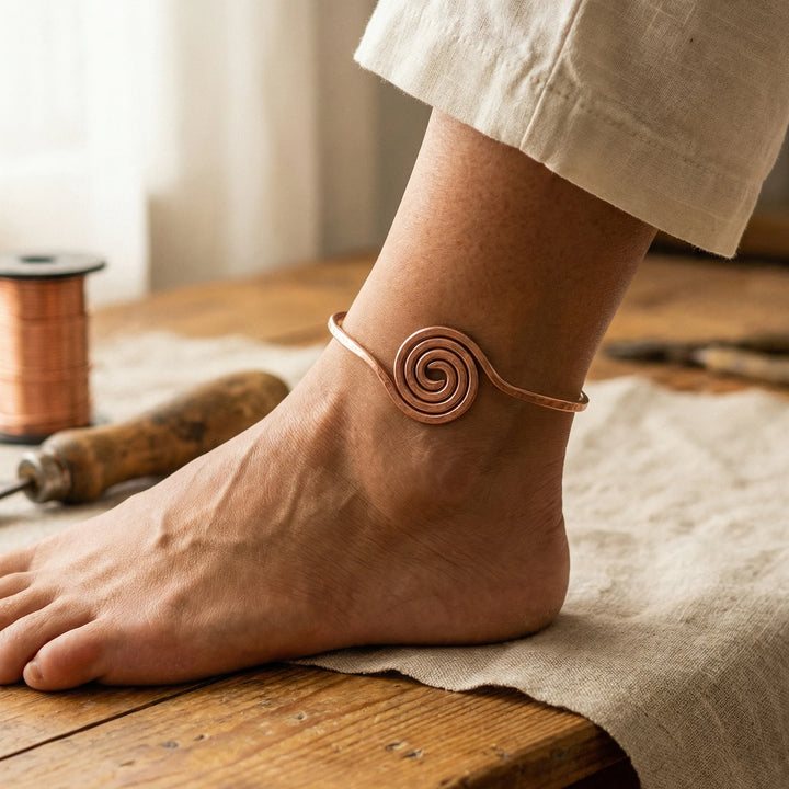 Copper spiral anklet worn around ankle with beige linen trousers on wooden floor